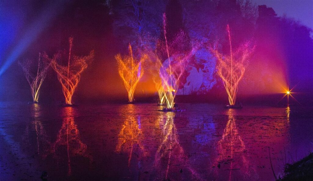 Trees illuminated in purple, pink and golden light reflected in the lake at Leonardslee Lights.