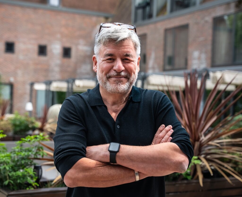 Grey-haired man in a black shirt standing outdoors with his arms folded, pictured for the Midhurst Jazz, Food & Blues Festival.