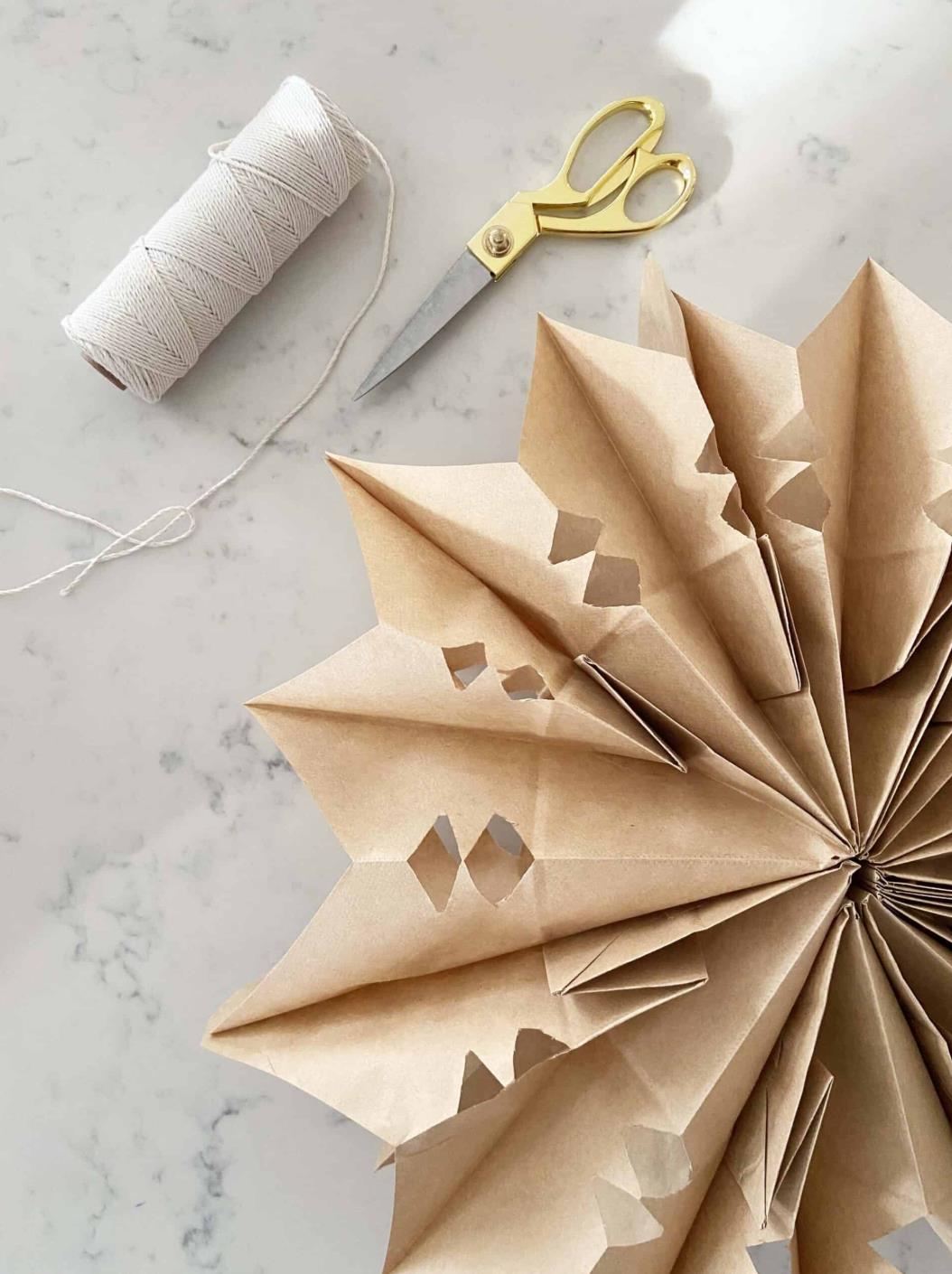 Folded brown paper star decoration beside gold-handled scissors and a spool of white string on a pale surface.