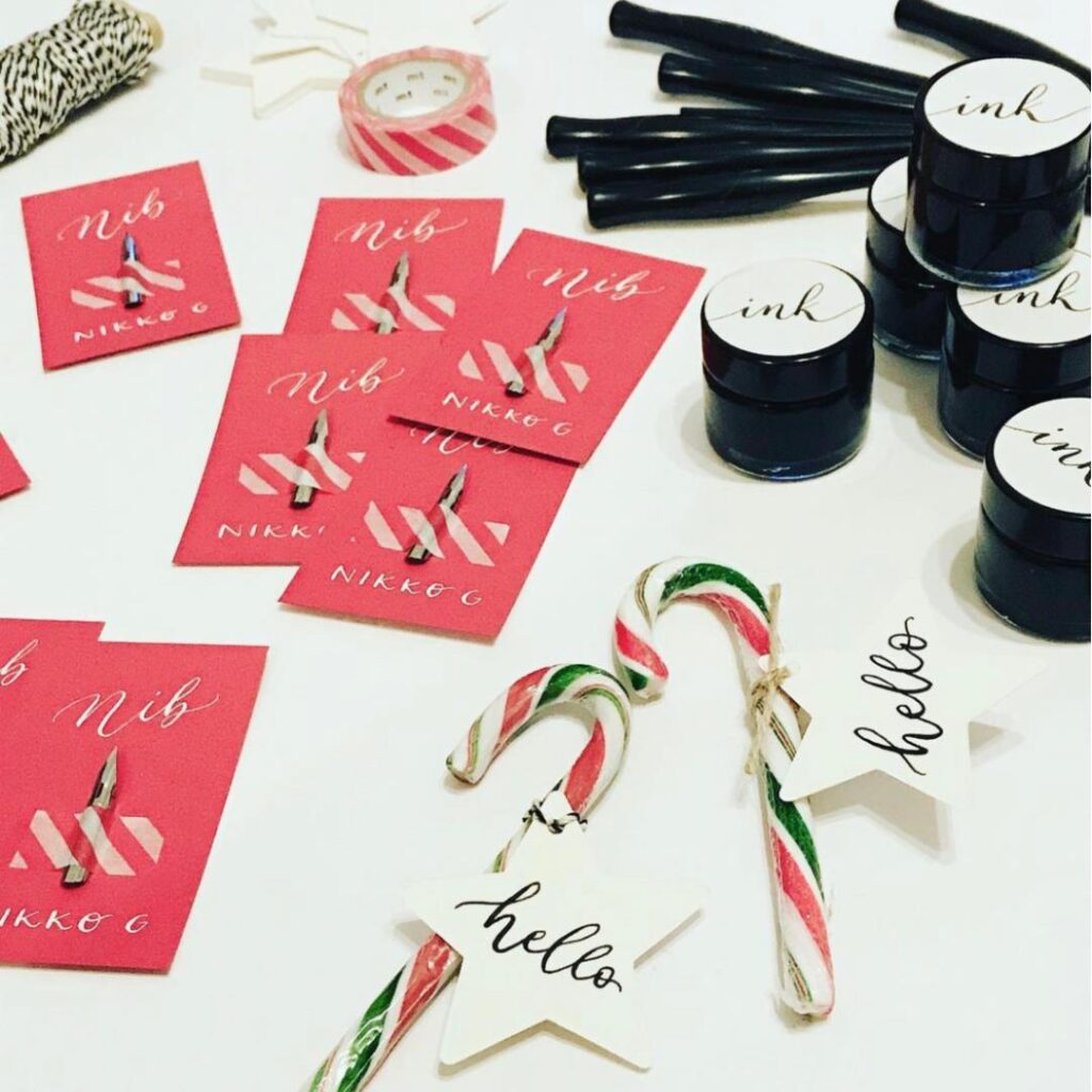 Dip pen nibs, black ink pots and pen holders beside candy canes and star-shaped tags with handwritten calligraphy.
