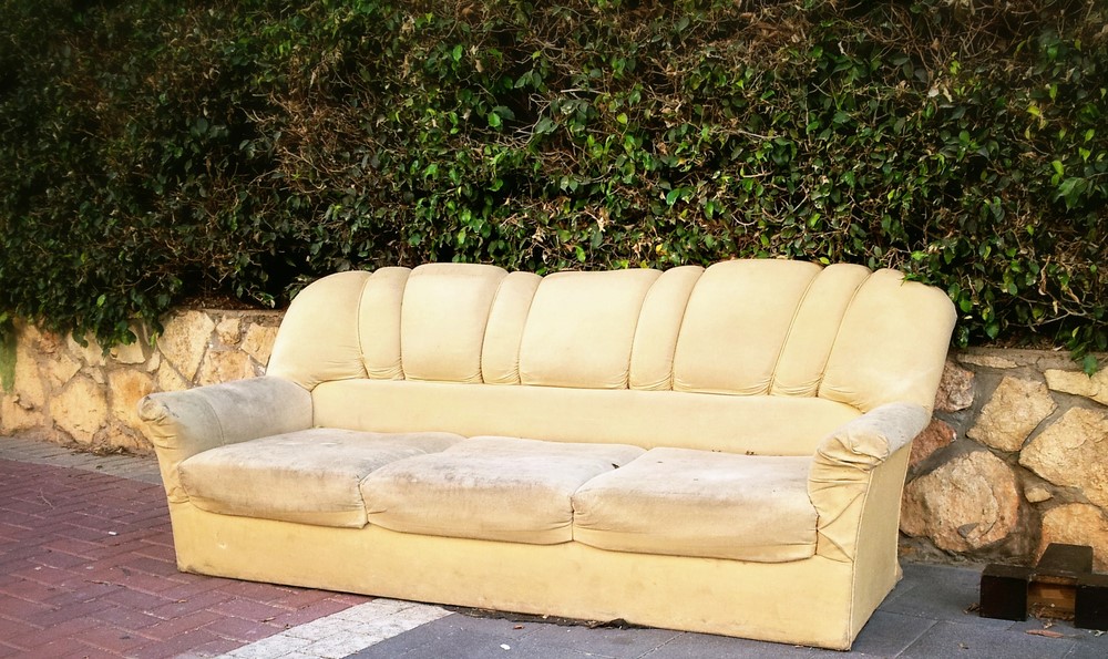 An old cream sofa on the pavement beside a stone wall and hedge