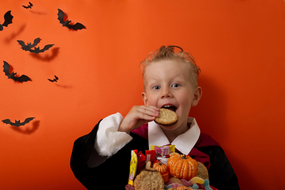 Child in a vampire Halloween costume eating a biscuit with sweets and small pumpkins against an orange backdrop with bats