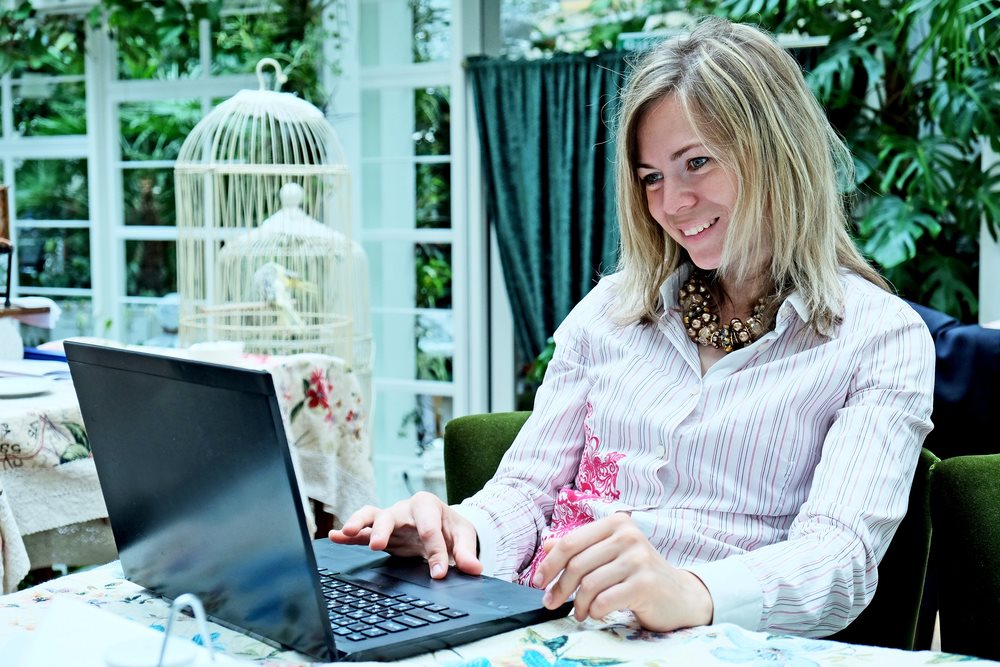 Smiling woman working on a laptop at a table in a bright conservatory surrounded by greenery.