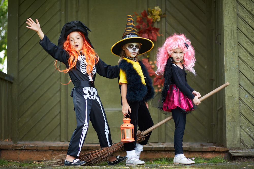 Three children wearing colourful Halloween costumes with wigs, a witch’s hat and a broomstick