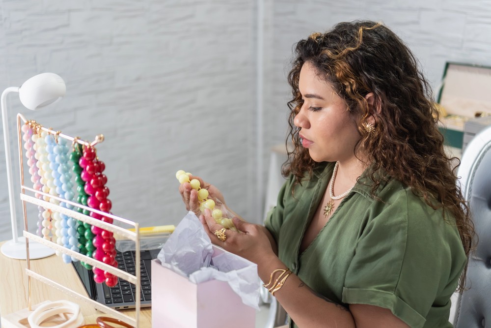 A woman holding colourful necklaces beside her laptop and jewellery packaging