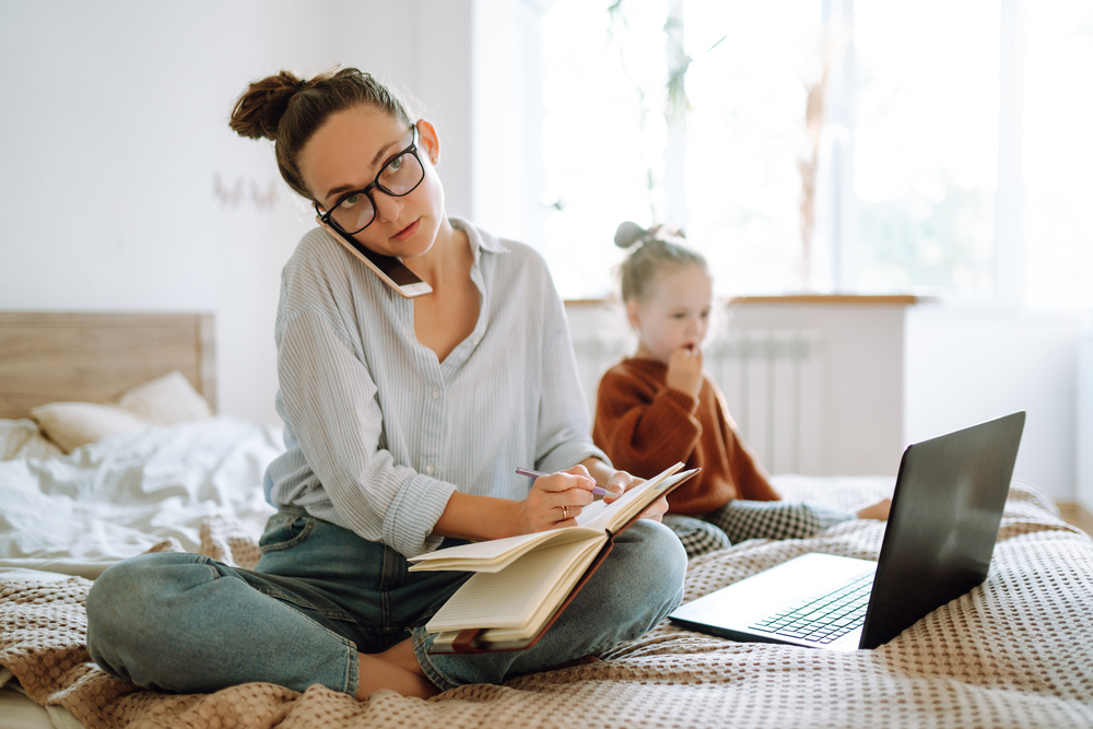Mother talking on the phone and writing in a notebook beside a laptop, with her child nearby.